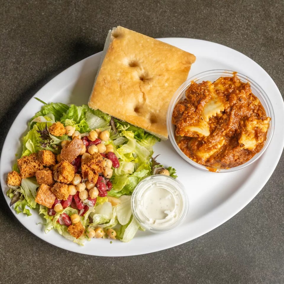 A white plate with a side salad, focaccia bread, ravioli in red sauce, and a cup of dressing.