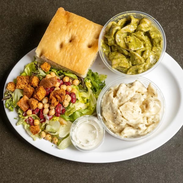 A white plate holds a focaccia bread, a salad, pesto pasta, potato salad, and dressing.