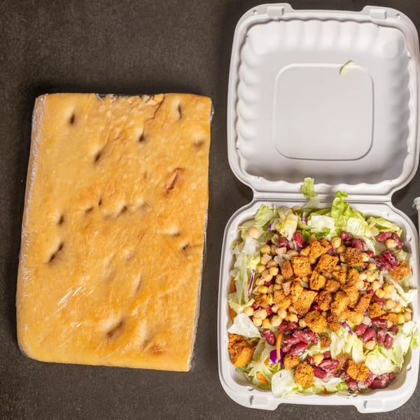Focaccia bread wrapped in plastic next to an open takeout container of salad with chickpeas and croutons.